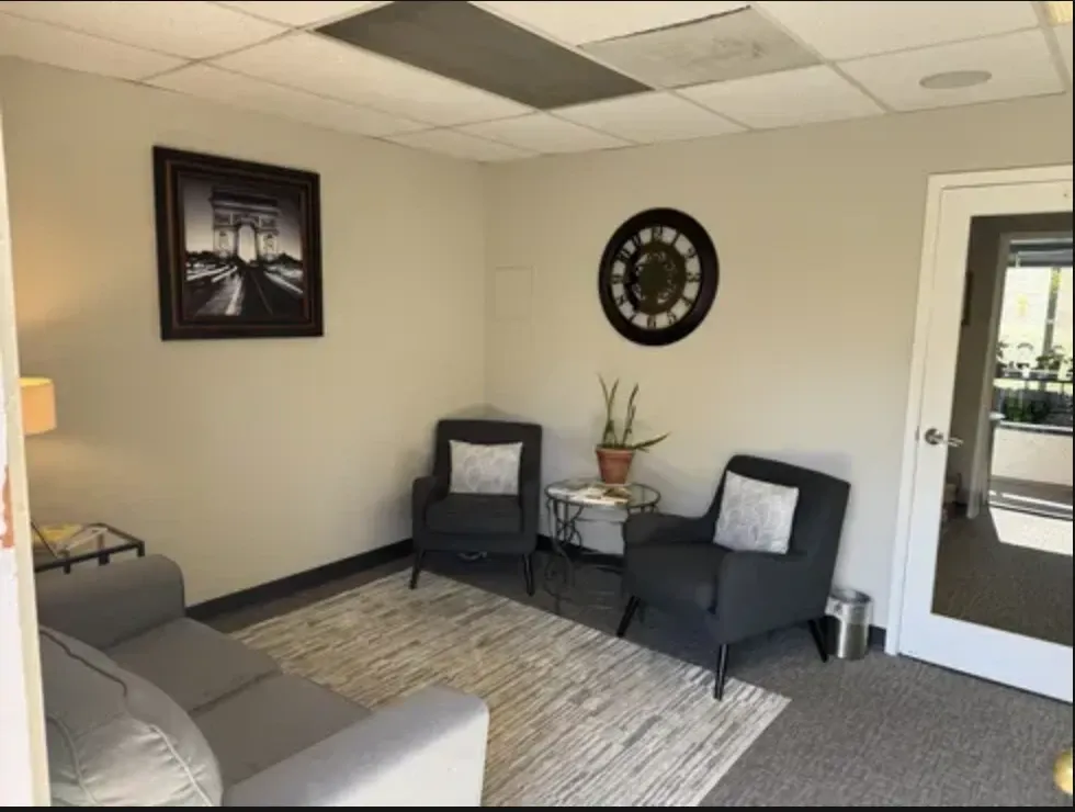 The office: two charcoal armchairs with white cushions beside a small round table and a plant, under a wall clock.